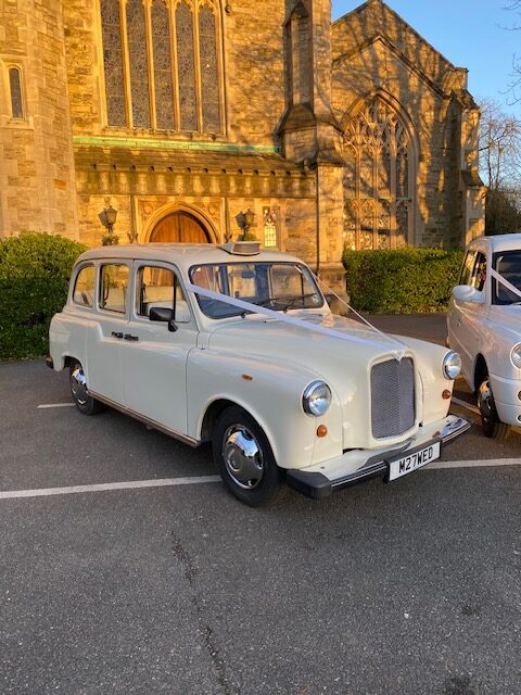 Classic Fairway wedding taxi with ivory ribbons parked outside a stone church